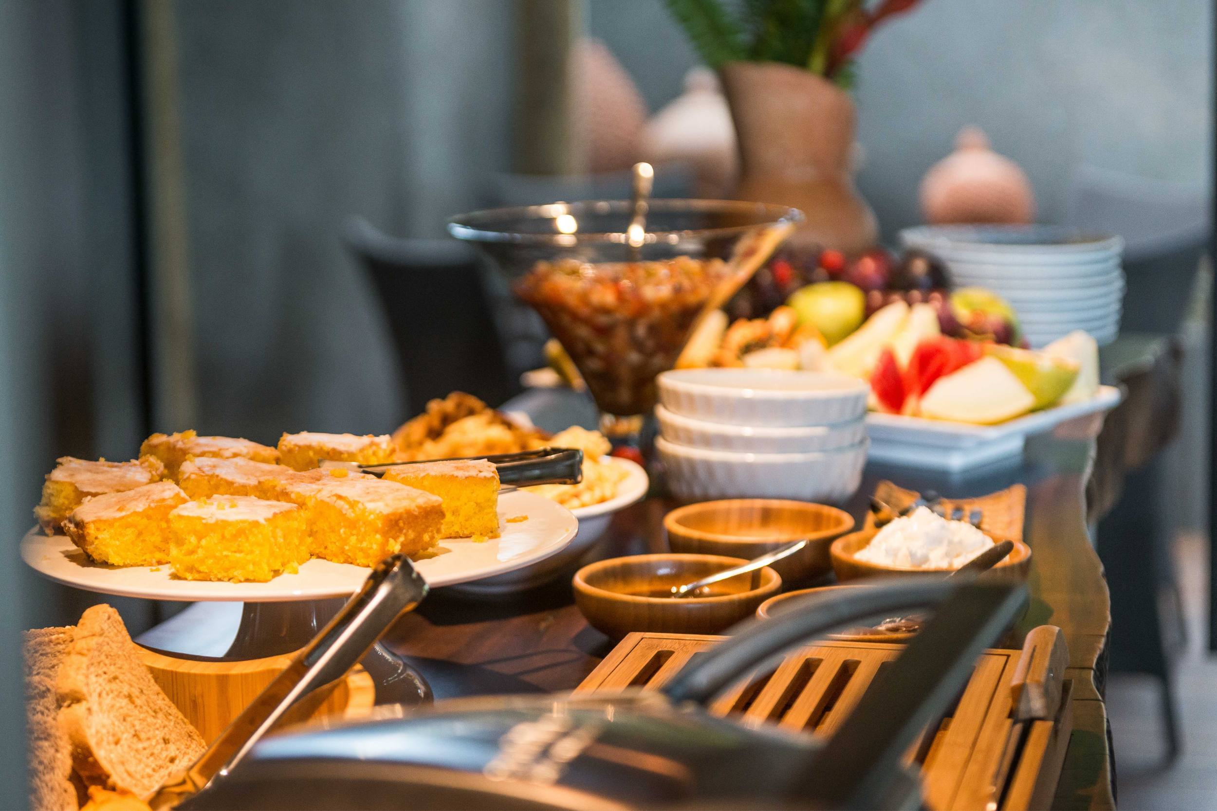 Mesa com produtos para café da manhã, entre os quais bolos, frios e frutas.
