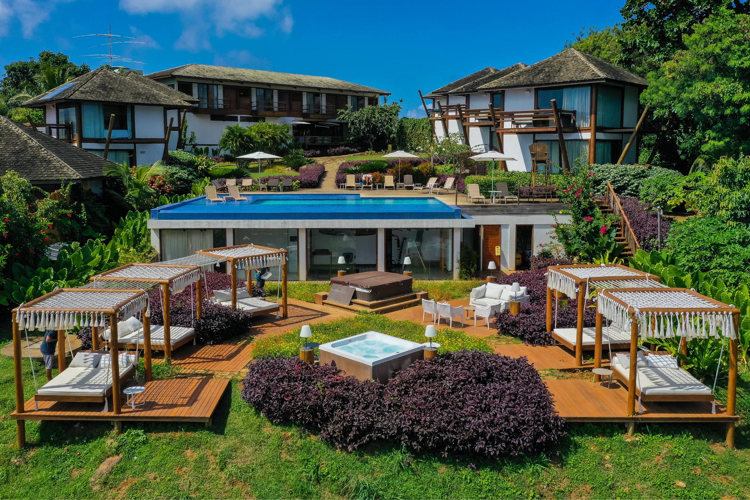 Lounge da pousada morena, com amplas espreguiçadeiras com dossel e banheira de hidromassagem. Acima, a piscina de borda infinita e o bloco central da pousada são visíveis.