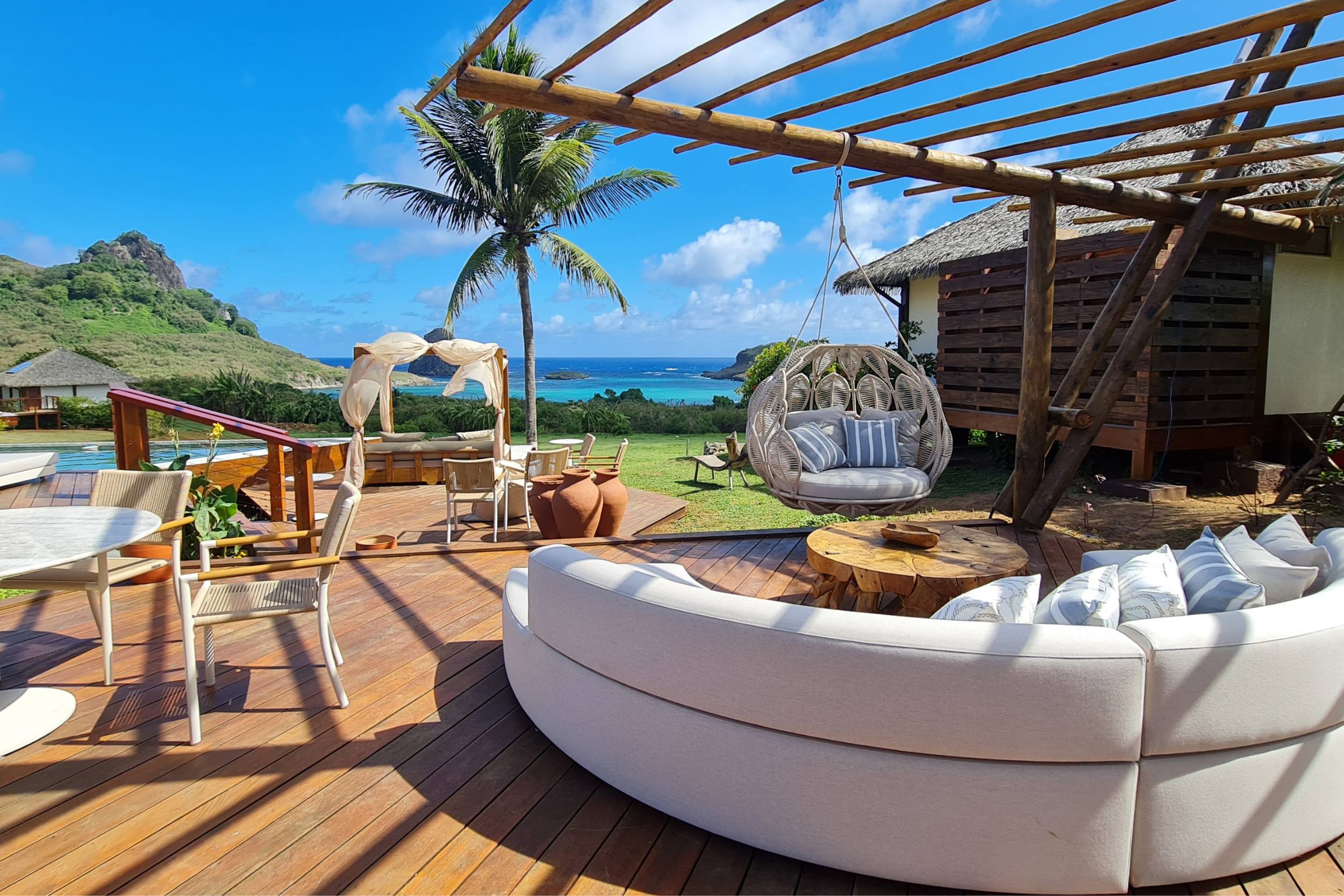 Um grande sofá estofado em formato semicircular sob um pergolado de madeira, com vista para a piscina da pousada e para o mar.